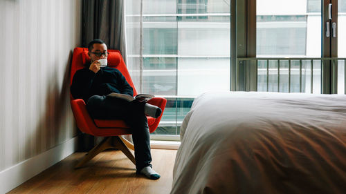 Full length of man drinking coffee while sitting at bedroom
