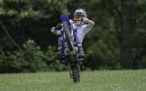 View of a young man riding a wheelie on a motorcycle in a field approaching with trees in background