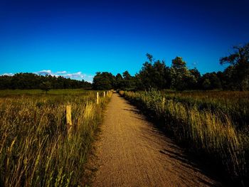 Dirt road amidst field against clear blue sky