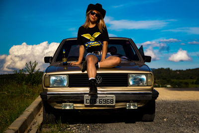 Portrait of young woman standing by car against sky