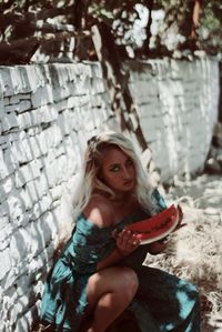 High angle portrait of young woman holding watermelon while crouching against wall