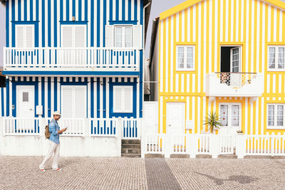 Side view of stylish man in hat using smartphone against creative facades of costa nova buildings