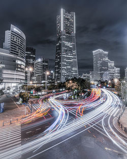 Light trails on city street by buildings against sky at night