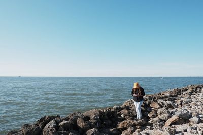 Rear view of woman sitting on rock at beach against clear sky