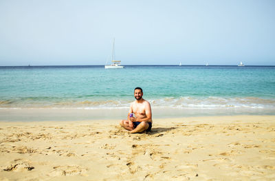 Portrait of shirtless mid adult man sitting on beach against clear sky during sunny day