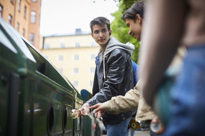 Teenage boy looking at friend while throwing waste in garbage bin at recycling station