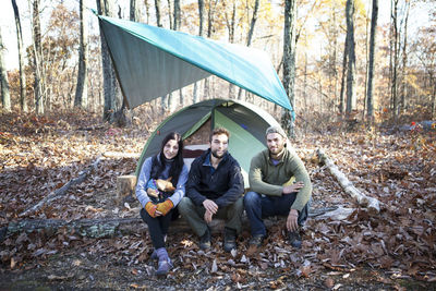 Portrait of friends sitting on fallen tree trunk by tent in forest