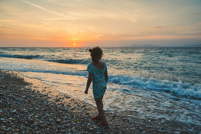 Rear view of girl standing on beach against sky during sunset