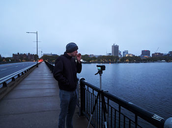 Man standing on railing by river against sky in city