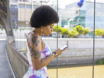 Side view of african american female with afro hairstyle and in fancy outfit standing on bridge in city and reading messages on social media via cellphone