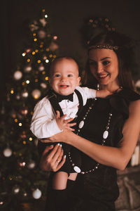 Mother carrying son while standing against christmas tree