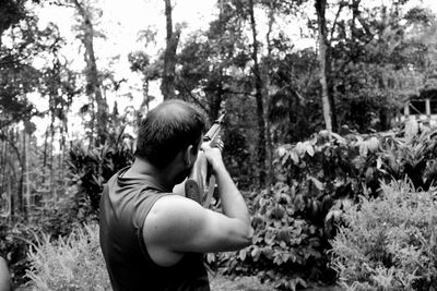 Man aiming rifle while standing against trees