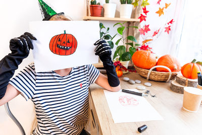 A child shows his drawing for halloween, sitting at a desk by the window, surrounded by pumpkins.