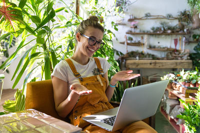 Smiling woman video conferencing over laptop while sitting amidst plants at home