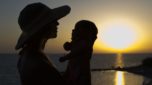 Mother carrying daughter at beach against sky during sunset