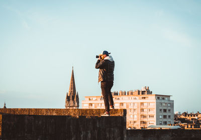 Man photographing while standing on retaining wall against clear sky