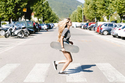 Young blond woman holding skateboard while running on zebra crossing in city