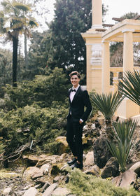 A beautiful young man, the groom in an elegant wedding suit, stands posing in the city's old park