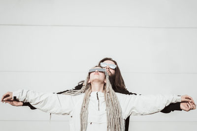 Young couple wearing sunglasses with arms outstretched standing against wall