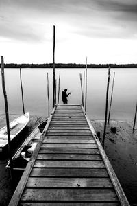 Rear view of man fishing in lake by pier against sky