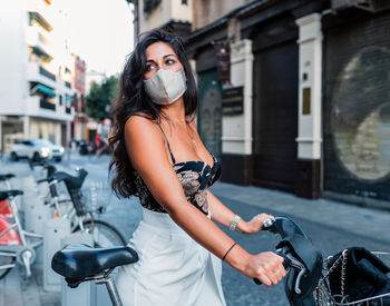 Side view of charming young latin female in stylish outfit and protective mask standing near bicycle on city street and looking away