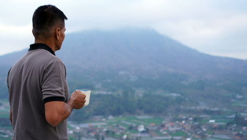 Side view of man looking at mountain with hand holding coffee or tea cup.