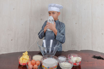 Portrait of boy wearing chef hat with ingredients on table standing against wall at home