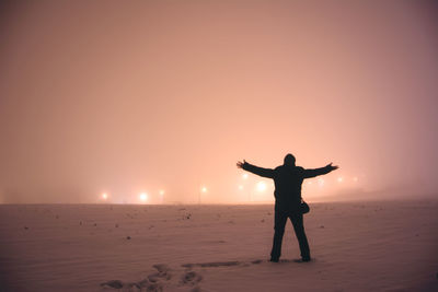 Rear view of man with arms outstretched standing on snow covered field at night