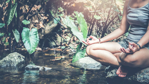 Low section of woman doing yoga while sitting on rock in forest