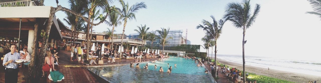 water, sea, beach, shore, nautical vessel, sand, clear sky, vacations, palm tree, large group of people, horizon over water, tree, boat, transportation, built structure, moored, day, sunlight, lounge chair