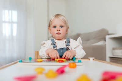 Charming little kid with blond hair assembling game pieces at table while looking at camera in house