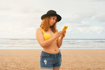 Caucasian redhead woman in a yellow bikini, hat and shorts with her phone standing on the beach
