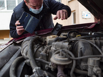 Midsection of man holding smart phone while repairing car engine