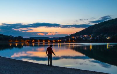 Rear view of silhouette man standing at lakeshore against sky during sunset