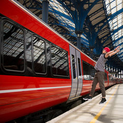 Full length of man with arms outstretched jumping by red train at railroad station