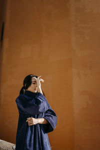 Young woman with eyes closed and head in hands in front of orange wall on sunny day