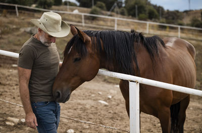 Full length of adult man with cowboy hat standing with brown horse on field