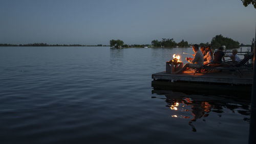 People sitting by lake against sky during sunset