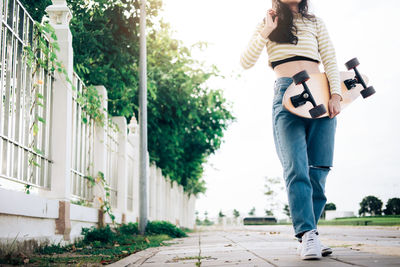 Low section of woman holding skateboard while walking on footpath