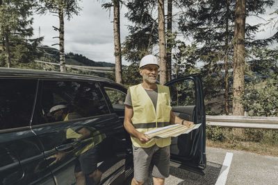 Smiling engineer holding plan standing by car