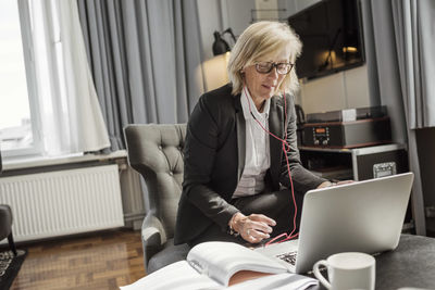 Mature businesswoman listening to headphones while using laptop in hotel room