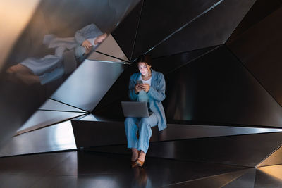 Full body of female in elegant wear text messaging on cellphone while sitting with netbook in modern studio with 3d walls