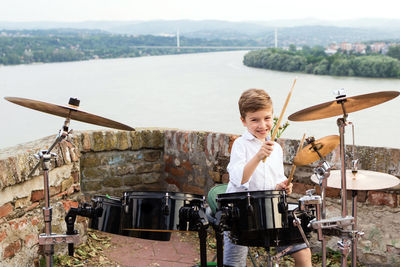 Smiling boy playing drum kit against river