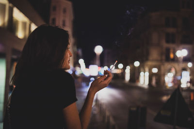 Side view of woman smoking cigarette while sitting in city at night