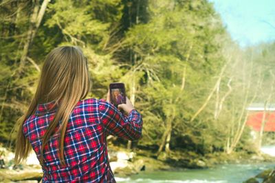 Rear view of woman taking selfie through mobile phone by stream at forest
