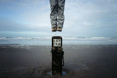 Low section of man levitating over wooden post at beach against cloudy sky