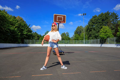 Full length of confident teenage girl holding ball while standing at basketball court during sunny day