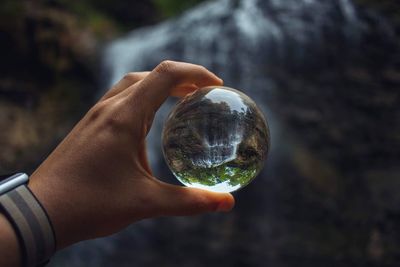 Cropped hand holding crystal ball with reflection against waterfall