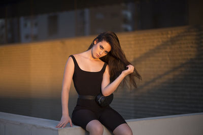 Serious young female in black bodysuit sitting on edge of border and looking at camera