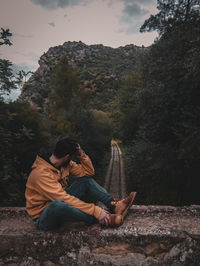 Full length of man sitting on stone wall while looking at railroad track